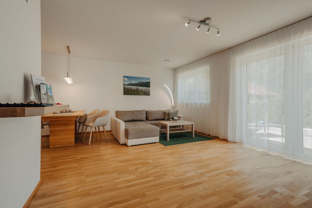 Blick vom Flur in helle Wohnküche mit Holzboden, Holztisch, hellen Stühlen und weiß grauem Sofa und Fensterfront mit Balkontür