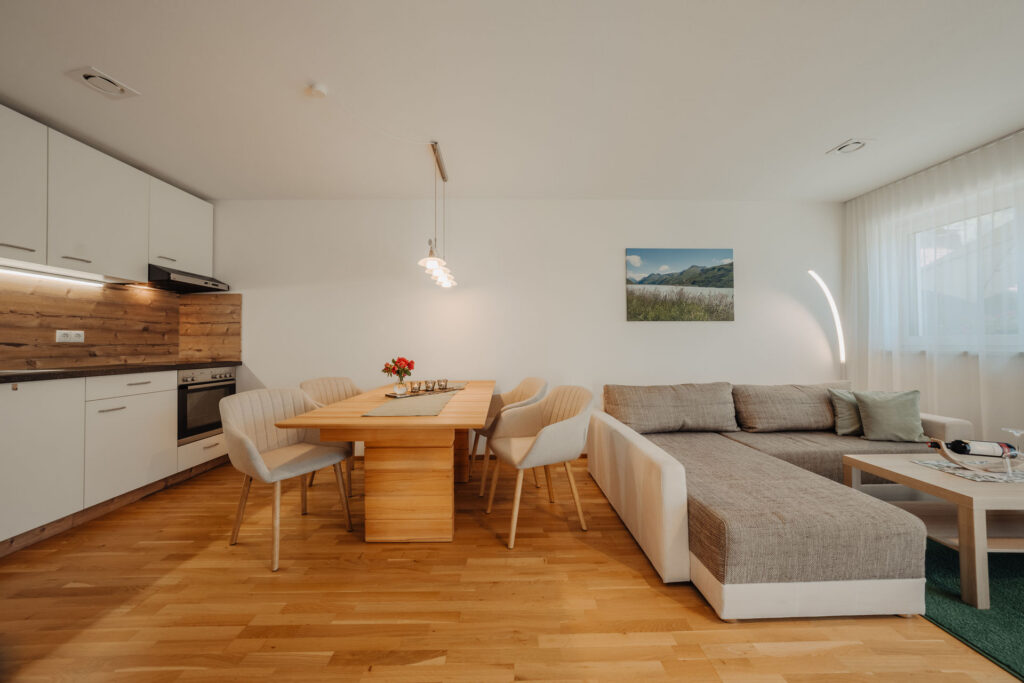 Blick vom Flur in helle Wohnküche mit Holzboden, Holztisch, hellen Stühlen und weiß grauem Sofa